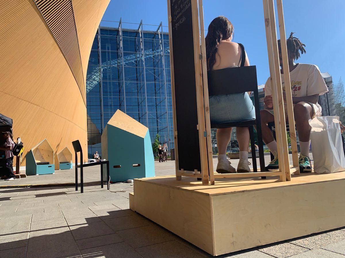 Two people sitting facing each other on a wooden platform. Their faces are not visible. The arc of Oodi library is in the background.