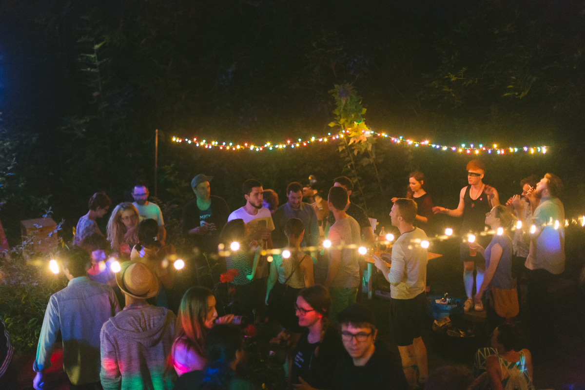 A party of around 20 people in a garden at evening time, talking in groups. Two colorful light strings hanging above them.